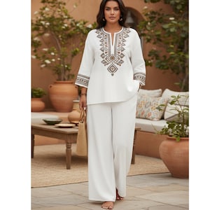 Puede incluir: Un conjunto de lino blanco con una túnica con bordados intrincados alrededor del cuello y las mangas, combinado con pantalones de pernera ancha. La parte superior tiene cuello en V y mangas tres cuartos. Se sostiene un bolso tote de paja en la mano.
