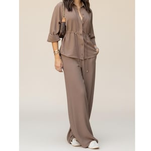 Puede incluir: Un conjunto de dos piezas color taupe con una camisa abotonada con cintura con cordón y pantalones de pernera ancha. La camisa tiene mangas enrolladas. El conjunto se combina con zapatillas blancas y un pequeño bolso marrón con una cadena dorada.