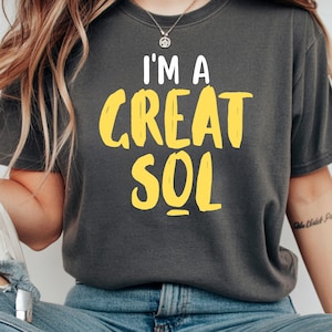 Può includere: T-shirt grigio scuro con la scritta "I'M A GREAT SOL" in bianco e giallo. La maglietta è in stile casual, adatta all'uso quotidiano. La persona indossa jeans blu.