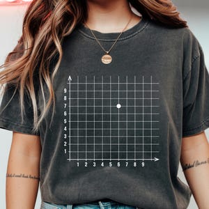 Puede incluir: Camiseta gris oscuro con un diseño de cuadrícula de coordenadas blanca y un punto en las coordenadas (6,7). La cuadrícula tiene ejes numerados del 1 al 9. Estilo casual y unisex.