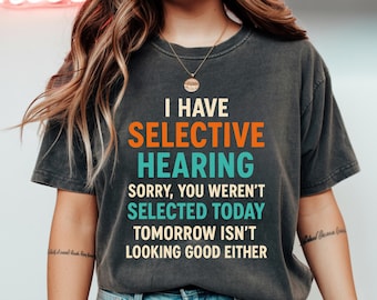 Może przedstawiać: Ciemnoszara koszulka z napisem "I HAVE SELECTIVE HEARING" w kolorze pomarańczowym i turkusowym. Koszulka kontynuuje frazami "SORRY, YOU WEREN'T SELECTED TODAY TOMORROW ISN'T LOOKING GOOD EITHER" w kolorze białym.