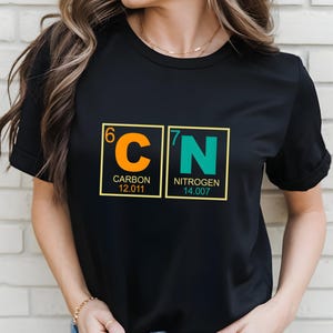 May include: Black t-shirt featuring a science-themed design. The graphic displays the chemical symbols for Carbon and Nitrogen, with atomic numbers and weights. The Carbon symbol is orange, and the Nitrogen symbol is teal.