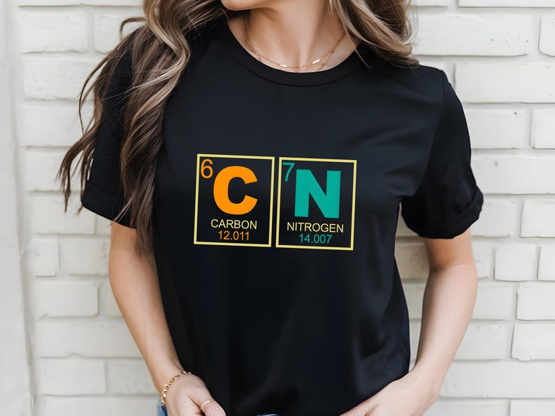 May include: Black t-shirt featuring a science-themed design. The graphic displays the chemical symbols for Carbon and Nitrogen, with atomic numbers and weights. The Carbon symbol is orange, and the Nitrogen symbol is teal.