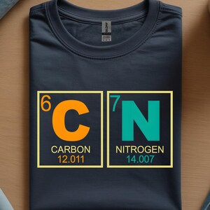May include: Navy blue t-shirt featuring the chemical symbols for Carbon and Nitrogen. The orange 'C' and teal 'N' are in yellow-bordered boxes, with the atomic numbers and weights below. A science-themed graphic tee.