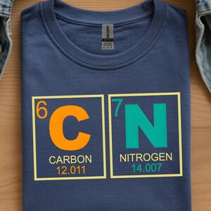 May include: Navy blue t-shirt featuring the chemical symbols 'C' and 'N' in orange and teal, representing Carbon and Nitrogen. The atomic numbers and weights are also displayed. A science-themed graphic tee.