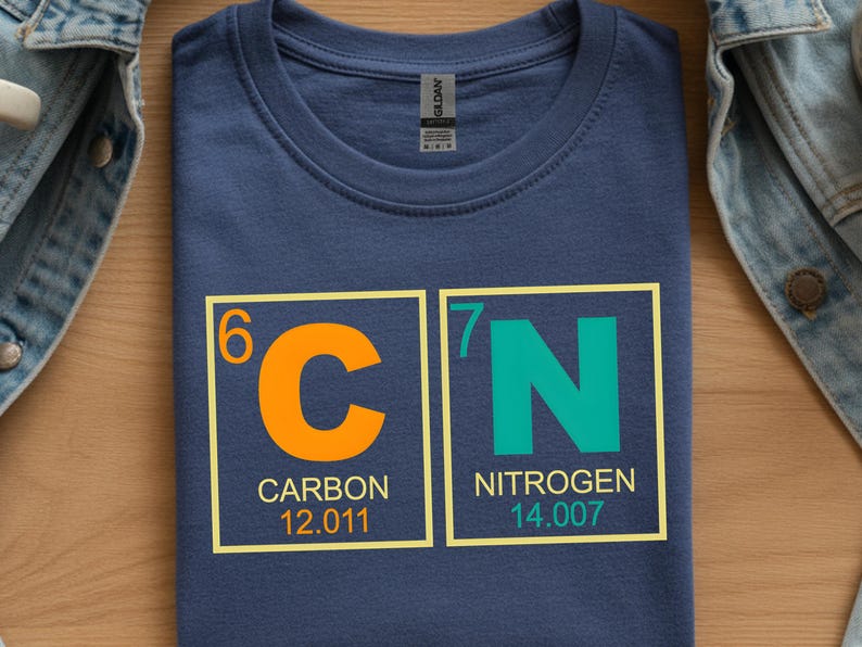 May include: Navy blue t-shirt featuring the chemical symbols 'C' and 'N' in orange and teal, representing Carbon and Nitrogen. The atomic numbers and weights are also displayed. A science-themed graphic tee.