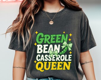 Puede incluir: Camiseta gris oscuro con el texto "Green Bean Casserole Queen" en verde y amarillo. El diseño incluye una vaina de judías verdes con un lazo y detalles de estrellas. Una divertida camiseta gráfica con temática de comida.