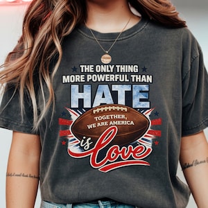 Può includere: T-shirt grigio scuro con la scritta "THE ONLY THING MORE POWERFUL THAN HATE IS LOVE". Il design include un pallone da football, elementi della bandiera americana e le parole "TOGETHER, WE ARE AMERICA."