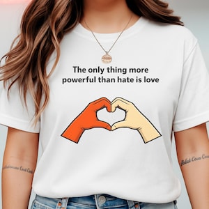 Könnte beinhalten: Weißes T-Shirt mit dem Text "The only thing more powerful than hate is love". Das Design zeigt zwei Hände, die ein Herz formen, eine orangefarbene und eine beige. Eine goldene Halskette ist sichtbar.