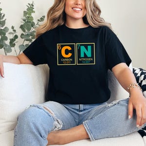May include: Black t-shirt featuring the chemical symbols for Carbon and Nitrogen, with atomic numbers and weights. The design is in orange and green, set against a yellow border. The person is wearing blue jeans.