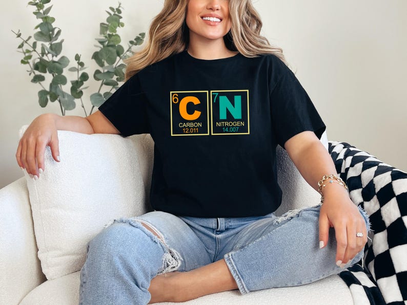 May include: Black t-shirt featuring the chemical symbols for Carbon and Nitrogen, with atomic numbers and weights. The design is in orange and green, set against a yellow border. The person is wearing blue jeans.