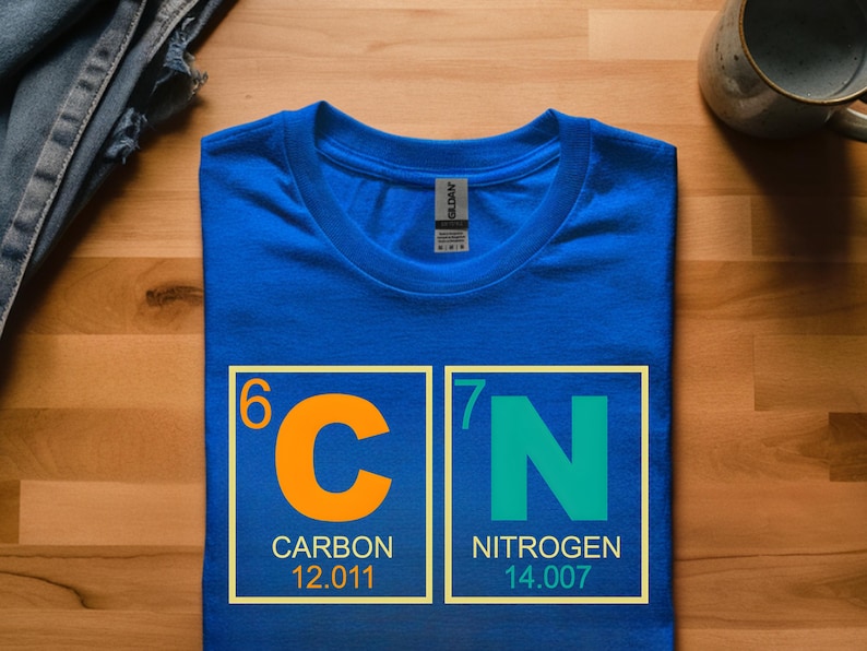 May include: A royal blue t-shirt featuring the periodic table symbols for Carbon and Nitrogen. The orange "C" is in a yellow box with the word "CARBON" and the number 12.011. The green "N" is in a yellow box with the word "NITROGEN" and the number 14.007.