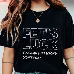 Puede incluir: Camiseta negra con las palabras "FET'S LUCK" en letras blancas grandes. Debajo, el texto dice "YOU READ THAT WRONG DIDN'T YOU?" en una fuente más pequeña. La camiseta es de estilo casual.