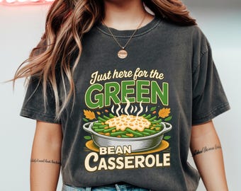 Puede incluir: Camiseta gris oscuro con un gráfico de cazuela de judías verdes. El texto dice "Just here for the GREEN Bean Casserole" en verde y amarillo. Un estilo de camisa casual y cómodo.