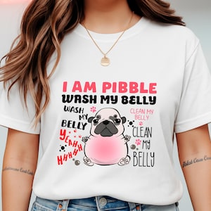 May include: White t-shirt featuring a cartoon pug with a pink belly and the text "I AM PIBBLE WASH MY BELLY." The design includes paw prints and additional text. A gold necklace is visible.