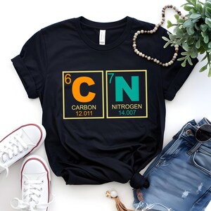 May include: Black t-shirt featuring the chemical symbols for Carbon and Nitrogen, with atomic numbers and weights. The shirt has a crew neck and is paired with white sneakers and blue jeans. A necklace and sunglasses are also visible.