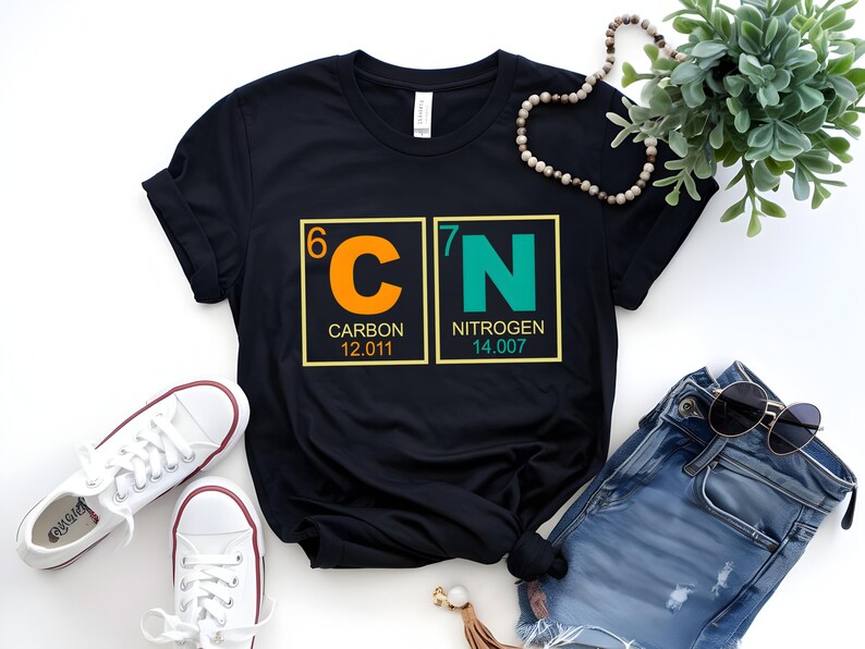 May include: Black t-shirt featuring the chemical symbols for Carbon and Nitrogen, with atomic numbers and weights. The shirt has a crew neck and is paired with white sneakers and blue jeans. A necklace and sunglasses are also visible.
