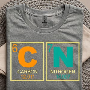 May include: A gray t-shirt featuring a design of the periodic table elements carbon and nitrogen. The carbon symbol is orange, and the nitrogen symbol is teal. The shirt includes the atomic numbers and weights for each element.