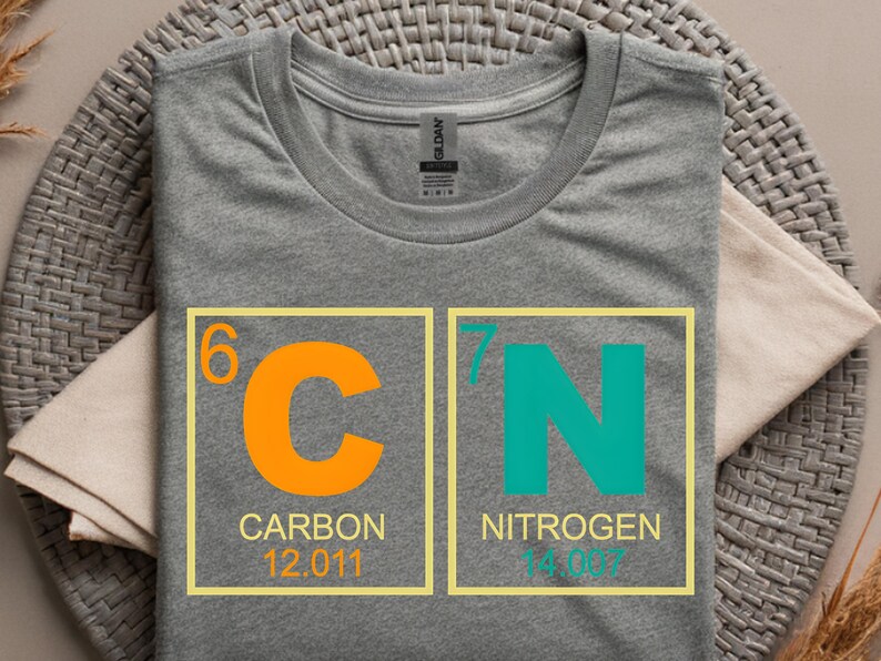 May include: A gray t-shirt featuring a design of the periodic table elements carbon and nitrogen. The carbon symbol is orange, and the nitrogen symbol is teal. The shirt includes the atomic numbers and weights for each element.