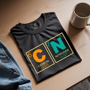 May include: Black t-shirt with a science-themed graphic featuring the chemical symbols for Carbon (C) and Nitrogen (N). The design includes the atomic numbers and weights. A beige mug sits nearby.