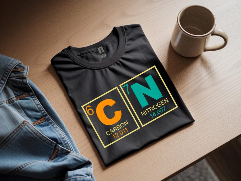 May include: Black t-shirt with a science-themed graphic featuring the chemical symbols for Carbon (C) and Nitrogen (N). The design includes the atomic numbers and weights. A beige mug sits nearby.