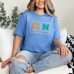May include: A light blue t-shirt with a graphic of the periodic table elements Carbon and Nitrogen. The shirt has the element symbols C and N in orange and green, with the atomic numbers and names below. The model is wearing ripped jeans.