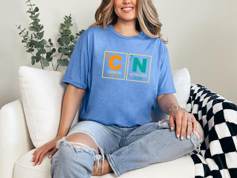 May include: A light blue t-shirt with a graphic of the periodic table elements Carbon and Nitrogen. The shirt has the element symbols C and N in orange and green, with the atomic numbers and names below. The model is wearing ripped jeans.