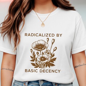 以下が含まれることがあります： 茶色の花の模様と「RADICALIZED BY BASIC DECENCY」の文字が入った白いTシャツ。このグラフィックは、花と種子の詳細な植物図を示しています。