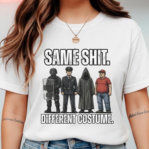 May include: White t-shirt with the text "SAME SHIT. DIFFERENT COSTUME." The graphic features four figures in different outfits: a riot officer, a police officer, a hooded figure, and a man in a red shirt and baseball cap.