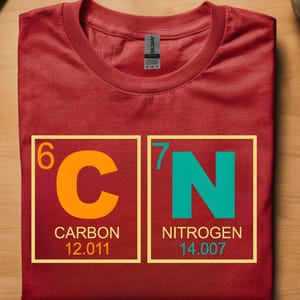 May include: A red t-shirt with a graphic of the periodic table elements carbon and nitrogen. The carbon symbol is orange, and the nitrogen symbol is teal. The shirt is folded on a wooden surface.