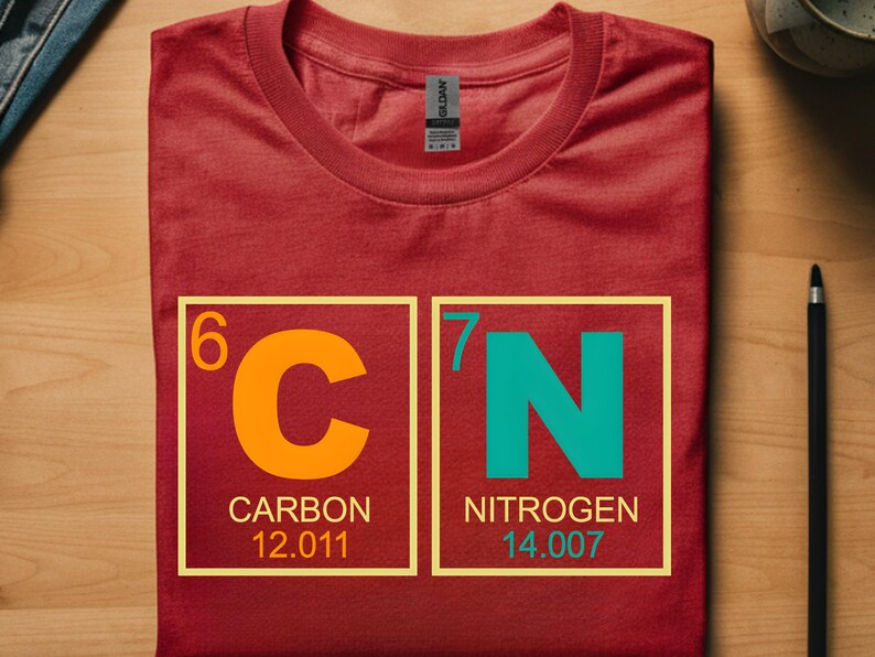 May include: A red t-shirt with a graphic of the periodic table elements carbon and nitrogen. The carbon symbol is orange, and the nitrogen symbol is teal. The shirt is folded on a wooden surface.