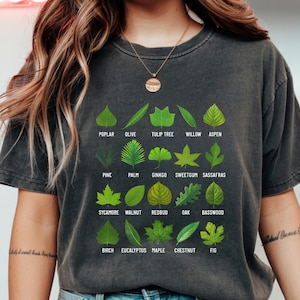 May include: Dark gray t-shirt featuring a botanical design of various leaf types, including poplar, olive, and willow. The names of the leaves are printed below each leaf. A stylish and educational tee for nature enthusiasts.