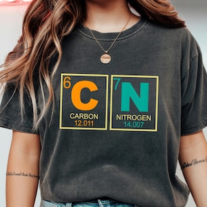 May include: Dark gray t-shirt featuring the chemical symbols for Carbon and Nitrogen. The orange and teal design includes the atomic numbers, element names, and atomic weights. The shirt is a comfortable, casual style.
