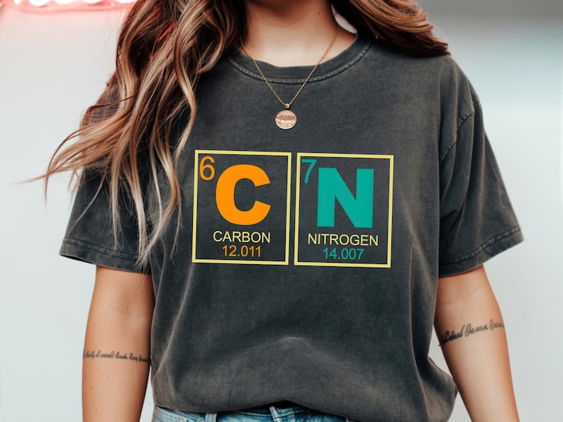 May include: Dark gray t-shirt featuring the chemical symbols for Carbon and Nitrogen. The orange and teal design includes the atomic numbers, element names, and atomic weights. The shirt is a comfortable, casual style.