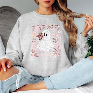 Könnte beinhalten: Hellgraues Sweatshirt mit einem weißen Geist, der Blumen hält, umgeben von einem rosa und braunen Blumendesign. Der Geist hat schwarze Augen. Das Sweatshirt ist ein lässiger, bequemer Stil.