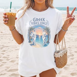 Könnte beinhalten: Ein weißes T-Shirt mit einem Grafikdruck und dem Text GREEK VIBES ONLY. Das Motiv zeigt zwei Figuren an einem Strand bei Sonnenuntergang, umrahmt von Olivenbäumen und einer blauen dekorativen Bordüre. Das T-Shirt hat kurze Ärmel und einen Rundhalsausschnitt.
