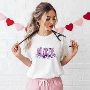 Könnte beinhalten: Weißes T-Shirt mit dem Wort "NURSE" in lila, floralem und kariertem Design. Das T-Shirt hat einen Rundhalsausschnitt und kurze Ärmel. Das Design umfasst lila Blumen und Blätter. Die Person trägt eine rosa Hose und hält ein Stethoskop.