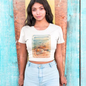 Könnte beinhalten: Ein weißes Crop-Top mit einem Strandmotiv, das Muscheln und Meereswellen zeigt. Das Model trägt hellblaue Jeansshorts. Der Hintergrund ist eine Holzwand mit blauer, gelber und brauner Farbe.
