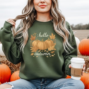 Könnte beinhalten: Ein dunkelgrünes Sweatshirt mit einem herbstlichen Motiv. Das Motiv zeigt drei orangefarbene Kürbisse, Herbstblätter und die Worte "hello autumn" in einer dekorativen Schriftart. Das Sweatshirt ist ein langärmeliger Rundhalsausschnitt.
