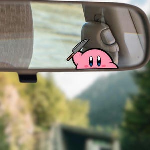 May include: A pink cartoon character with a knife sticker is placed on a car's rearview mirror. The character has large blue eyes and a smiling expression. The background shows a blurred view of a road and trees.