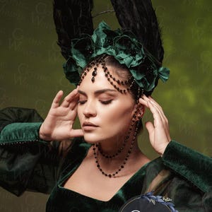 K&ouml;nnte beinhalten: Eine Frau in einem dunkelgr&uuml;nen Samtkleid mit einer schwarzen Perlenkette und einem dramatischen Kopfschmuck mit schwarzen Federn und gr&uuml;nen Samtblumen. Der Kopfschmuck ist mit schwarzen Perlen und einer Kette verziert. Der Hintergrund ist ein gr&uuml;ner Farbverlauf.