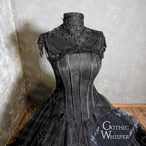 K&ouml;nnte beinhalten: Ein schwarzes Kleid im Gothic-Stil mit hohem Kragen und Korsett-Oberteil. Das Kleid hat aufwendige Spitzen- und Perlenverzierungen am Kragen und an den Schultern. Der volle Rock besteht aus T&uuml;llschichten. Der Text "Gothic Whisper" ist enthalten.