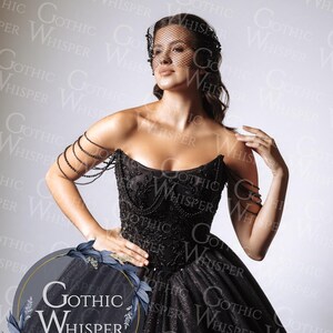 Op de afbeelding: Een zwarte strapless jurk met een geborduurd lijfje en een volle, sprankelende rok. De jurk heeft ingewikkeld kralenwerk en een decoratieve armband. Het model draagt een zwarte sluier. Het logo "Gothic Whisper" is zichtbaar.