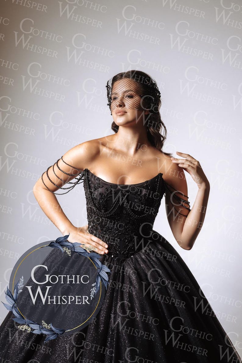 Op de afbeelding: Een zwarte strapless jurk met een geborduurd lijfje en een volle, sprankelende rok. De jurk heeft ingewikkeld kralenwerk en een decoratieve armband. Het model draagt een zwarte sluier. Het logo "Gothic Whisper" is zichtbaar.