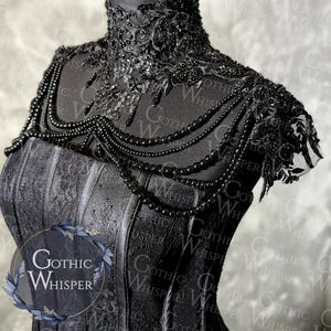 K&ouml;nnte beinhalten: Ein schwarzes Korsett mit aufwendigen Spitzendetails und einer Perlenkette. Das Korsett hat einen hohen, verzierten Kragen und ein strukturiertes Oberteil. Die Halskette f&auml;llt nach vorne und verleiht Eleganz. Das Gothic Whisper-Logo ist sichtbar.