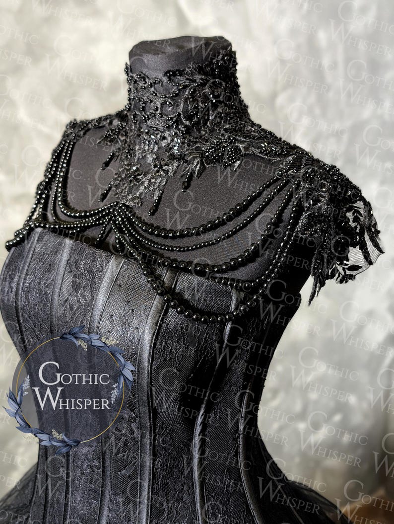 K&ouml;nnte beinhalten: Ein schwarzes Korsett mit aufwendigen Spitzendetails und einer Perlenkette. Das Korsett hat einen hohen, verzierten Kragen und ein strukturiertes Oberteil. Die Halskette f&auml;llt nach vorne und verleiht Eleganz. Das Gothic Whisper-Logo ist sichtbar.