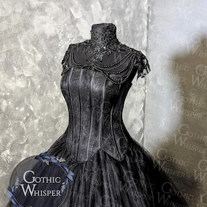 K&ouml;nnte beinhalten: Schwarzes Kleid im Gothic-Stil auf einer Schaufensterpuppe. Das Kleid hat ein detailliertes Oberteil mit vertikalen Linien und einen mehrlagigen T&uuml;llrock. Der Ausschnitt ist mit aufwendiger Spitze und Perlen verziert. Das Logo "Gothic Whisper" ist sichtbar.