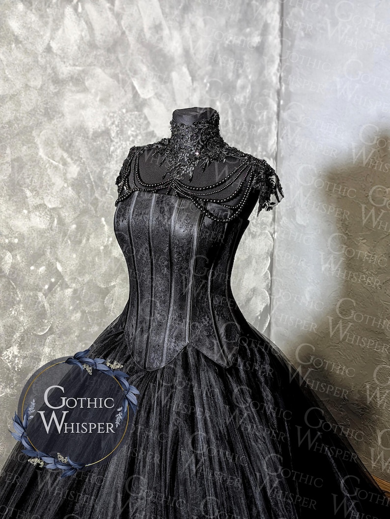 K&ouml;nnte beinhalten: Schwarzes Kleid im Gothic-Stil auf einer Schaufensterpuppe. Das Kleid hat ein detailliertes Oberteil mit vertikalen Linien und einen mehrlagigen T&uuml;llrock. Der Ausschnitt ist mit aufwendiger Spitze und Perlen verziert. Das Logo "Gothic Whisper" ist sichtbar.