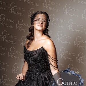Op de afbeelding: Een vrouw in een zwarte, geborduurde jurk met een tule sluier. De jurk heeft een sweetheart halslijn en een volle rok. De afbeelding bevat de tekst "Gothic Whisper" in een cirkelvormig logo. Het thema is gothic mode.