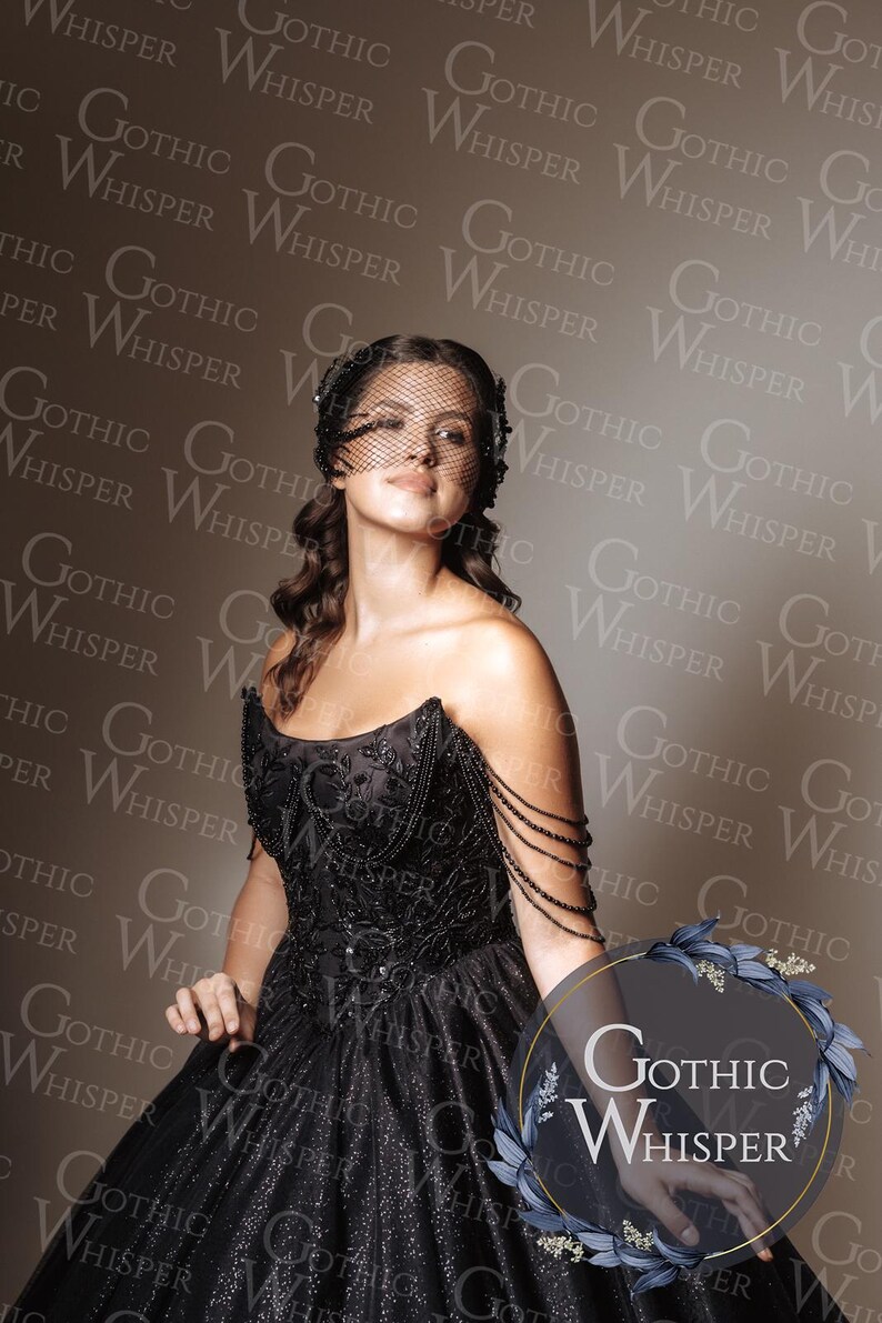Op de afbeelding: Een vrouw in een zwarte, geborduurde jurk met een tule sluier. De jurk heeft een sweetheart halslijn en een volle rok. De afbeelding bevat de tekst "Gothic Whisper" in een cirkelvormig logo. Het thema is gothic mode.